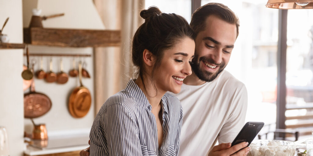 Photo of smiling brunette couple man and woman 20s drinking coffee and using cell phone during breakfast in kitchen at home