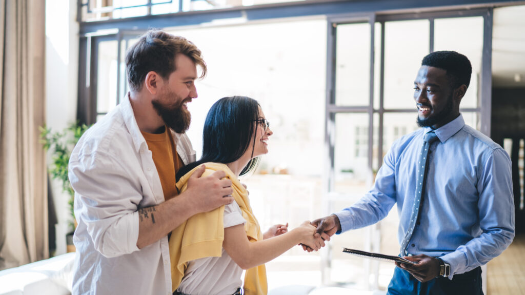 Bearded optimistic African American real estate broker greeting joyful couple by shaking hands with woman in glasses while standing in brightly lit apartment