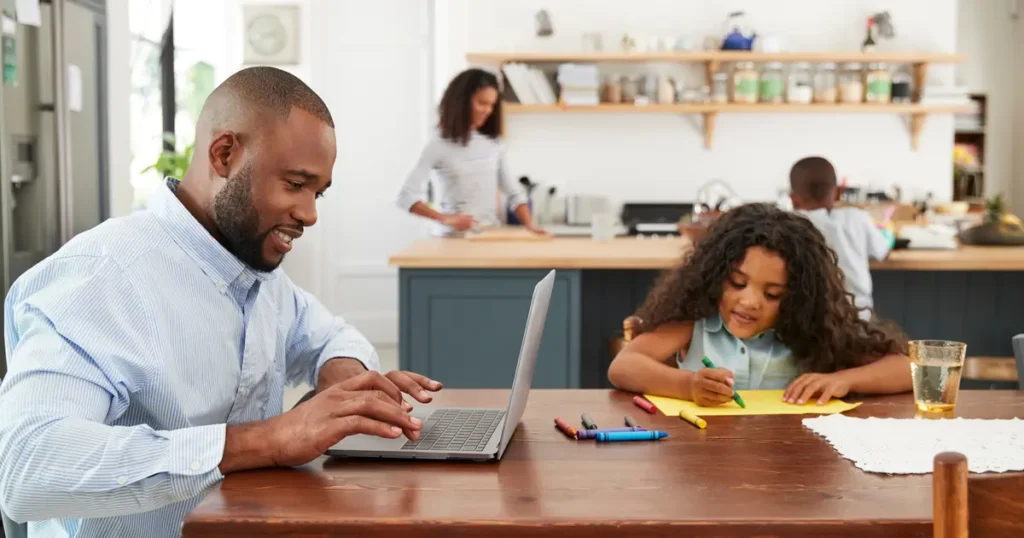A father working on a laptop while his daughter draws at the table, with the mother and son in the background of a bright kitchen." Let me know if you'd like to adjust this description!