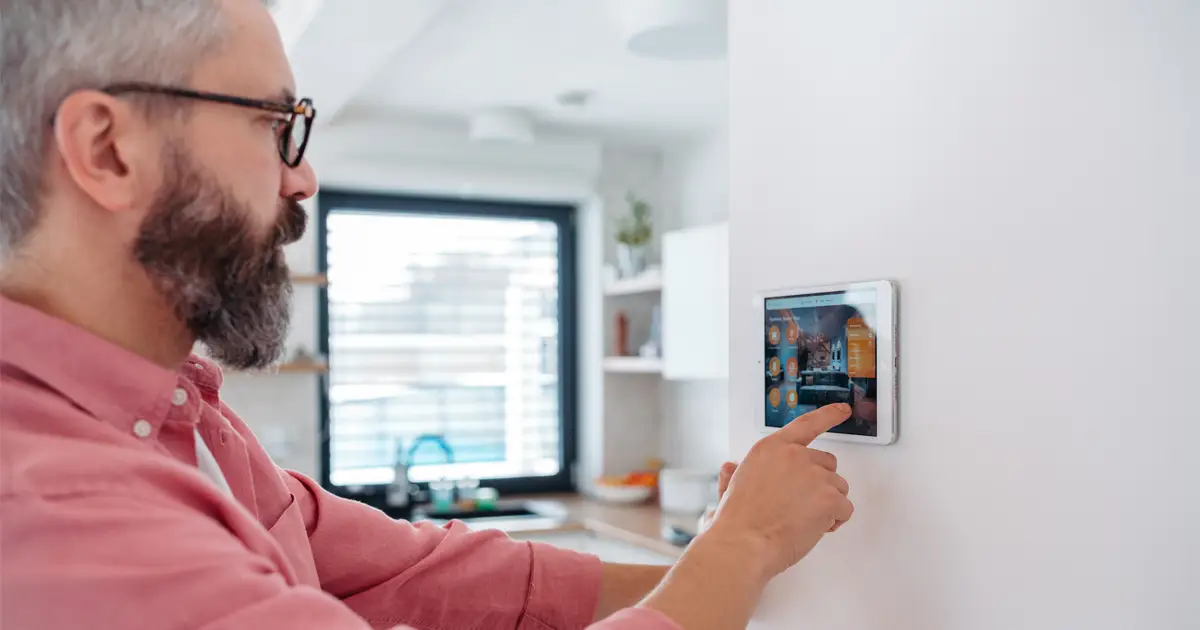 A bearded man wearing glasses and a pink button-up shirt interacts with a smart home control panel mounted on a white wall. The modern kitchen in the background has natural light coming through a window with blinds, along with shelves holding kitchen items and plants.