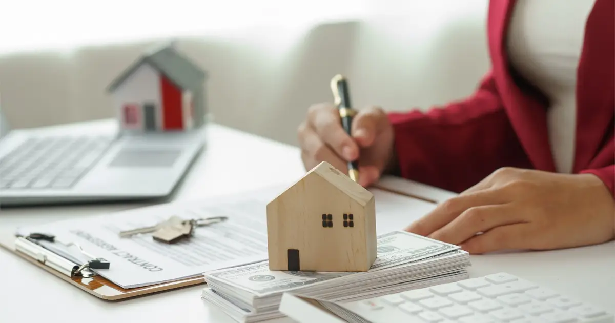 A person in a red blazer is signing a document at a desk, symbolizing a real estate or mortgage transaction. In the foreground, a small wooden house model sits atop a stack of cash, representing home financing. A clipboard with contract papers and house keys, a laptop, and another house model in the background complete the scene.