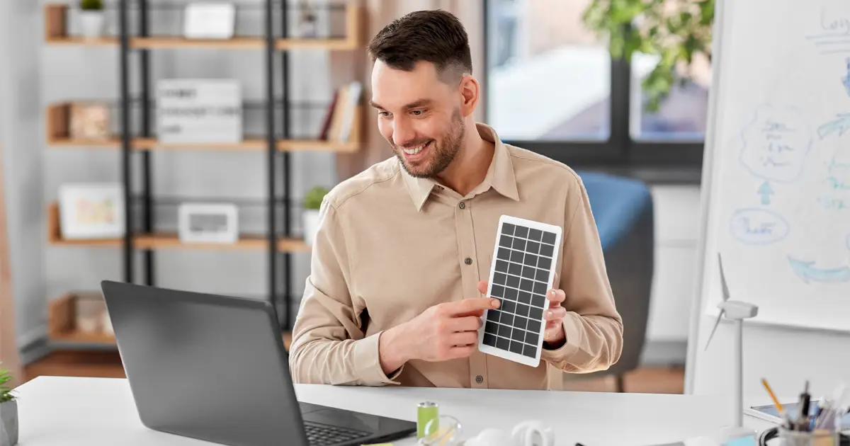 A smiling man in a beige shirt sits at a desk in a modern office, holding and pointing at a small solar panel. A laptop, light bulbs, a battery, and office supplies are on the desk. In the background, a whiteboard with diagrams and a small wind turbine model suggest a discussion about renewable energy.